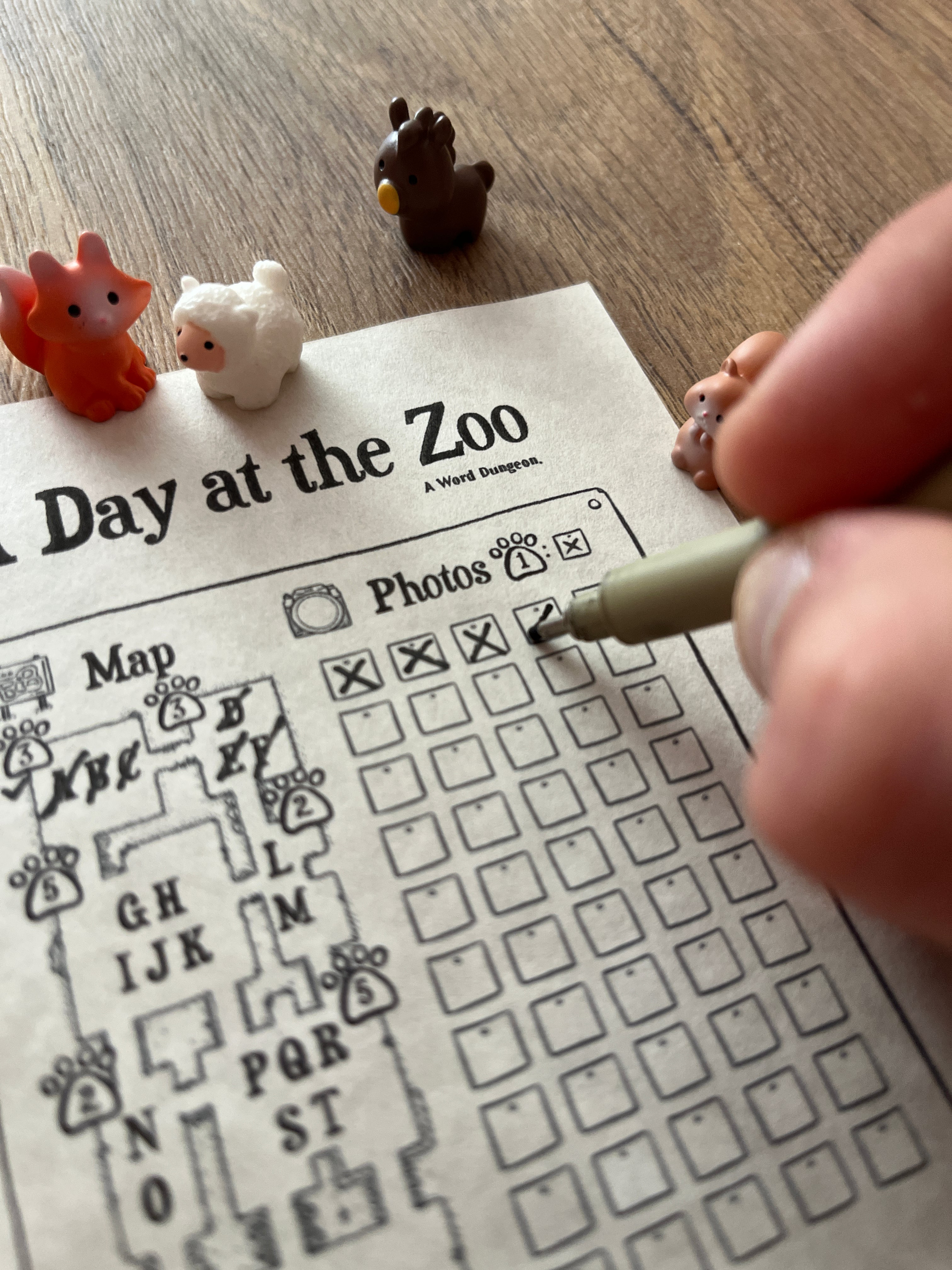 A close-up of a hand using a fine-liner pen to mark boxes on a printed zoo-themed activity sheet, with small animal figurines placed nearby on a wooden surface.